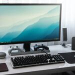 A computer monitor, keyboard, and game controllers on a white desk in a workspace