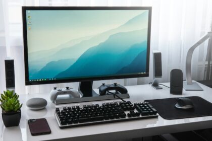 A computer monitor, keyboard, and game controllers on a white desk in a workspace