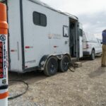 An orange fiber-optic cable warning marker beside a work trailer on a rural Utah site