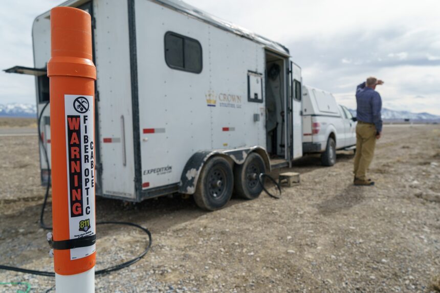 An orange fiber-optic cable warning marker beside a work trailer on a rural Utah site