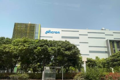 White Micron fab building with the Micron logo, behind a row of trees and a bus stop in Taichung, Taiwan