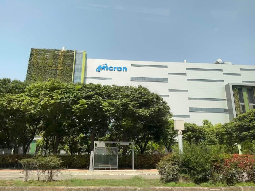 White Micron fab building with the Micron logo, behind a row of trees and a bus stop in Taichung, Taiwan